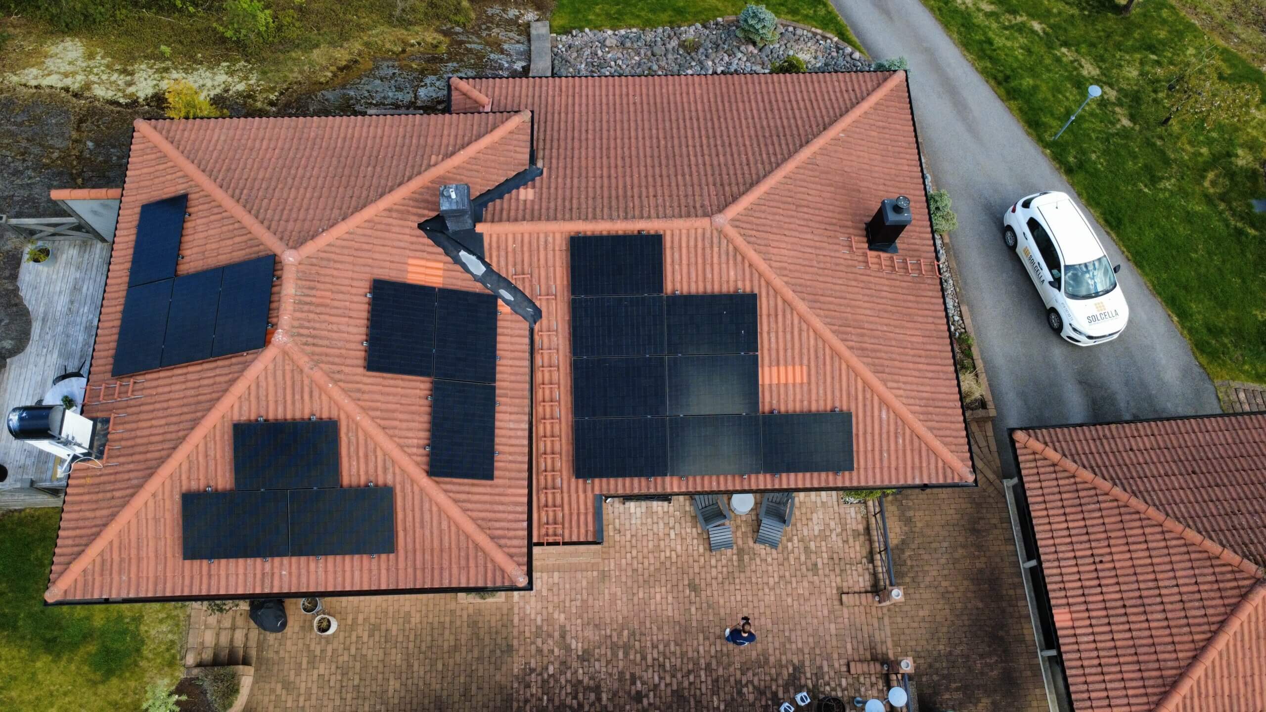 Solcellsanläggning på Värmdö Flygbild på ett brutet tegeltak på Värmdö med 18 nymonterade svarta solpaneler och en Solcella-bil parkerad på uppfarten.