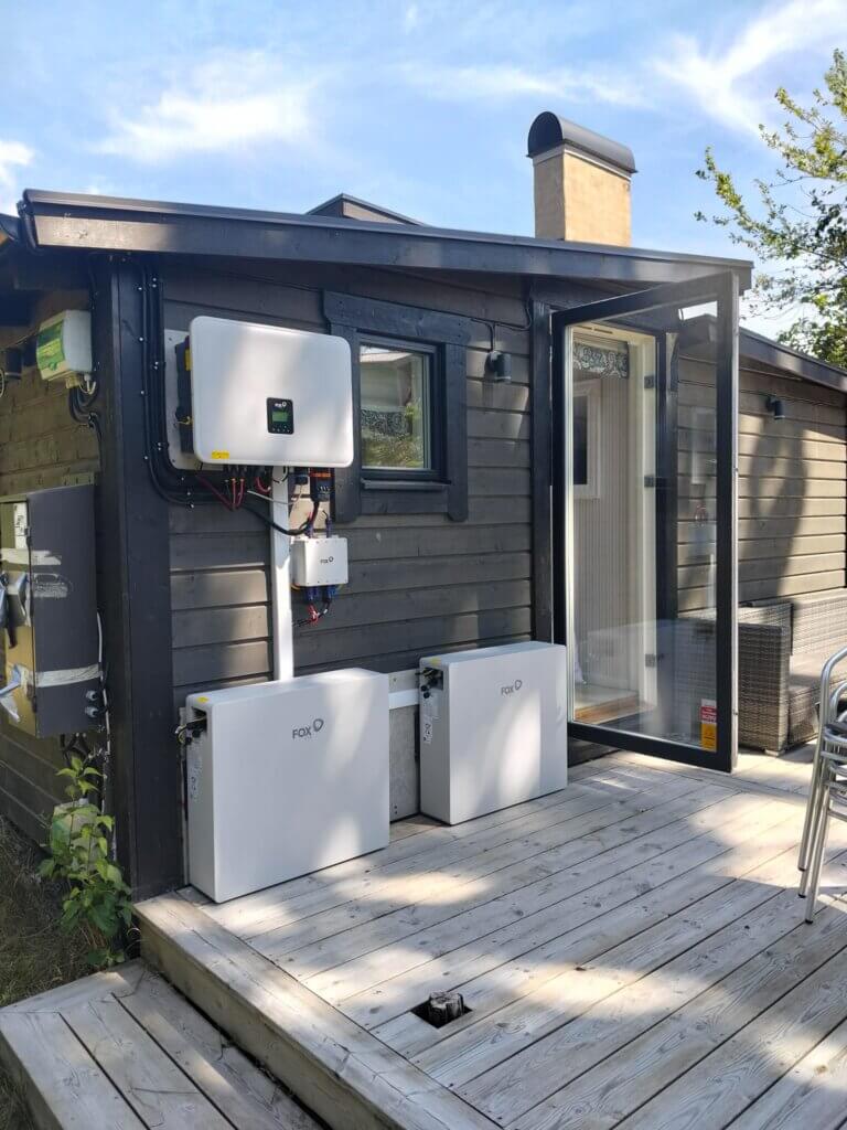 Utomhusinstallation på ett trädäck av ett stort 23 kWh batteri bestående av två moduler samt en växelriktare, båda från FoxESS, monterade på en mörk träfasad.