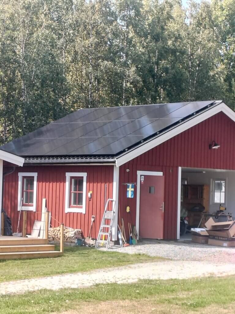 Rött garage med vita knutar där takytan har nyttjats maximalt med svarta solpaneler för högsta möjliga elproduktion.