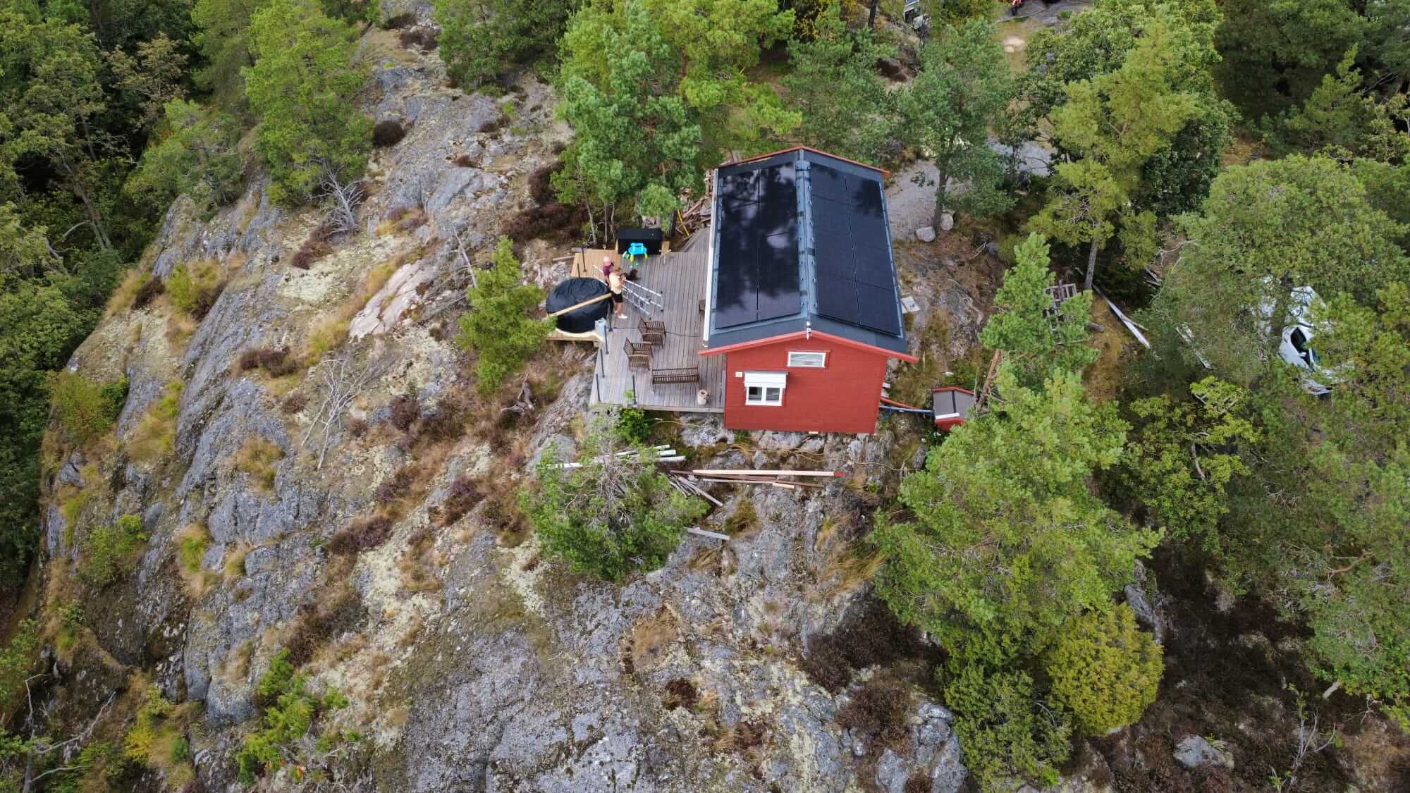 Utzoomad drönarbild över en röd sommarstuga högt uppe på en kulle på Bullandö, med nymonterade solceller på papptaket.