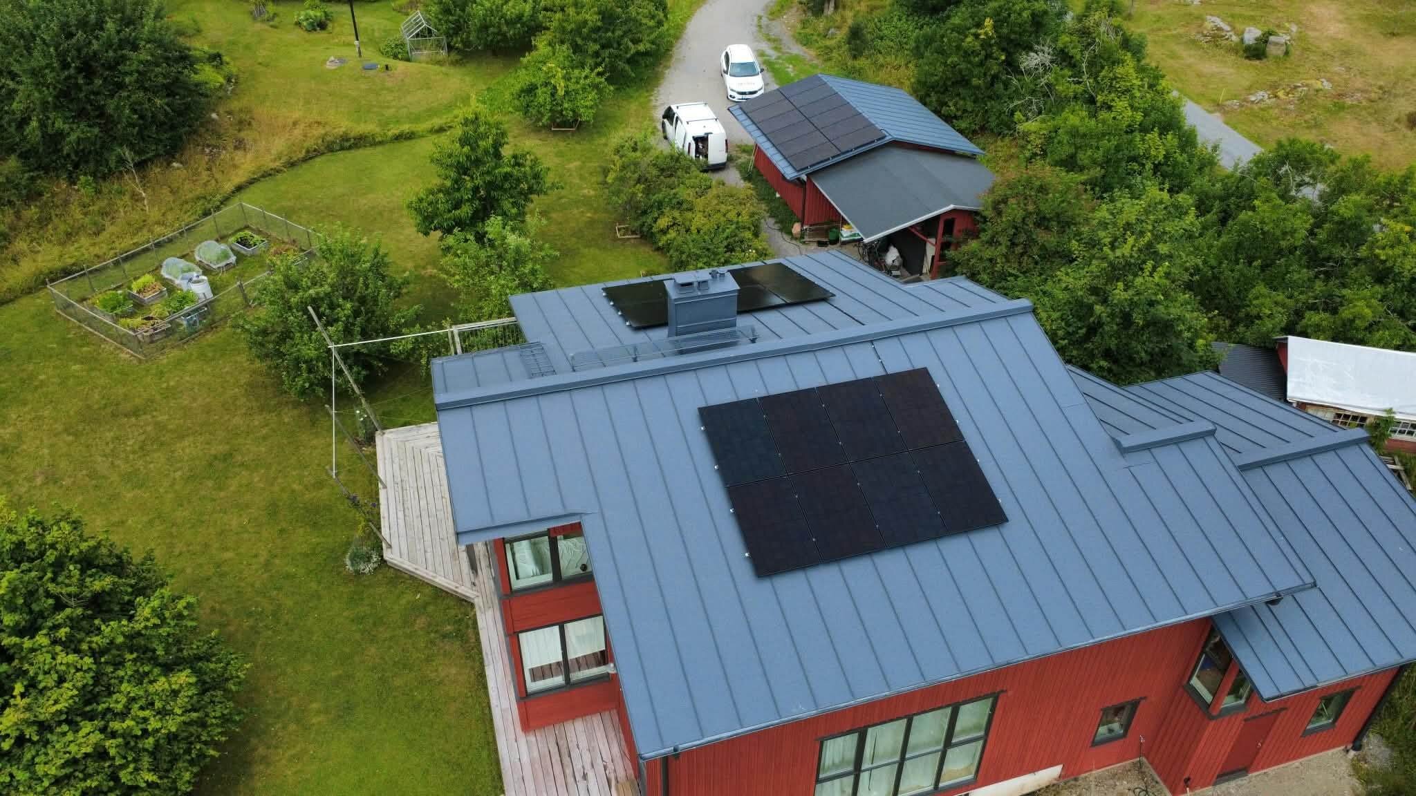 Drönarbild över en röd trävilla och ett garage på Ekerö med nyligen installerade solpaneler på plåttaken.